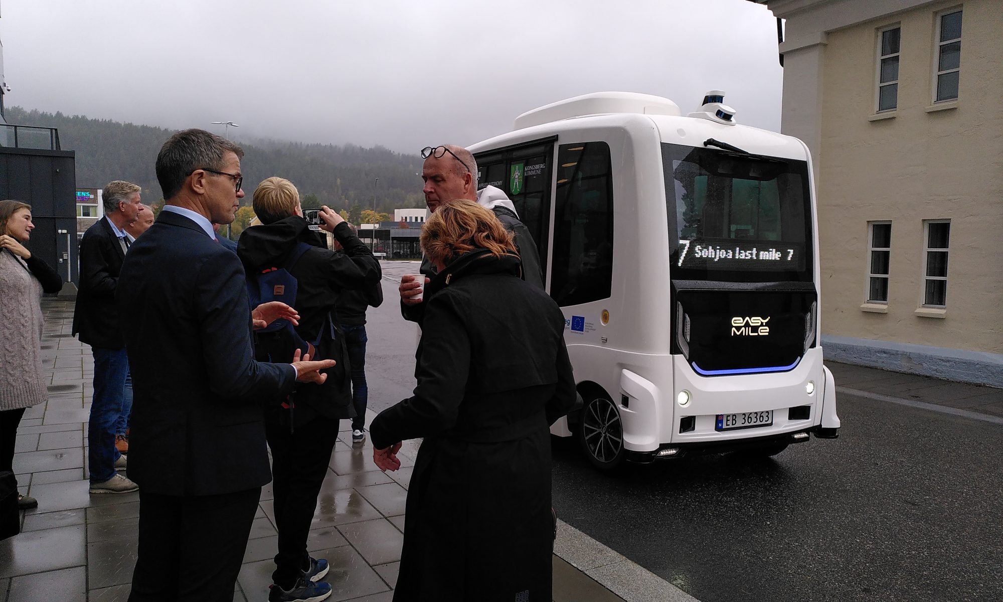 A group of people standing outside in the left, background grey and rainy skies and a silhouette of a hill, a robot bus on the road, a house on the right.